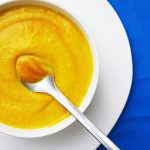 Carrot Soup in a white bow, with a spoon, on a white plate on a bright blue background