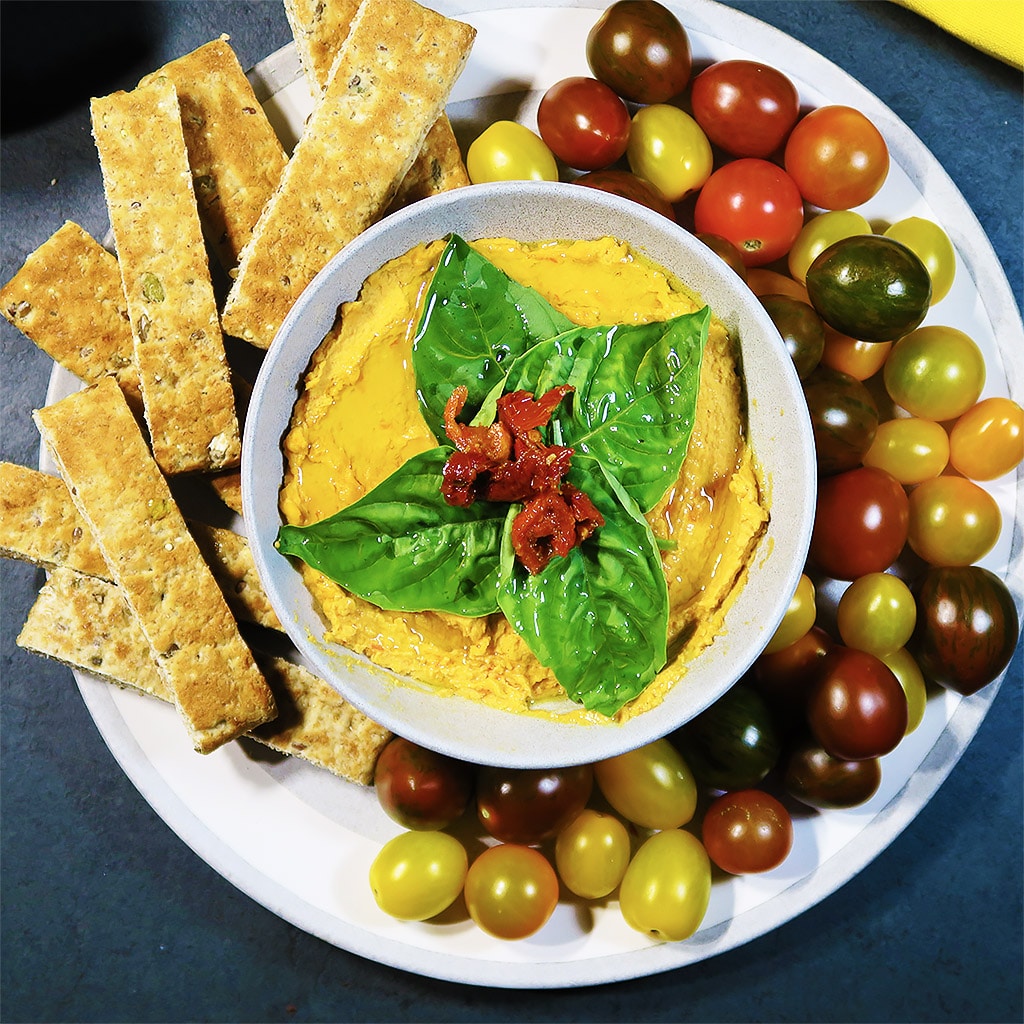 Top down view of 3-ingredient Sundried Tomato Hummus in a dish, surrounded by bread sticks and tomatoes. 