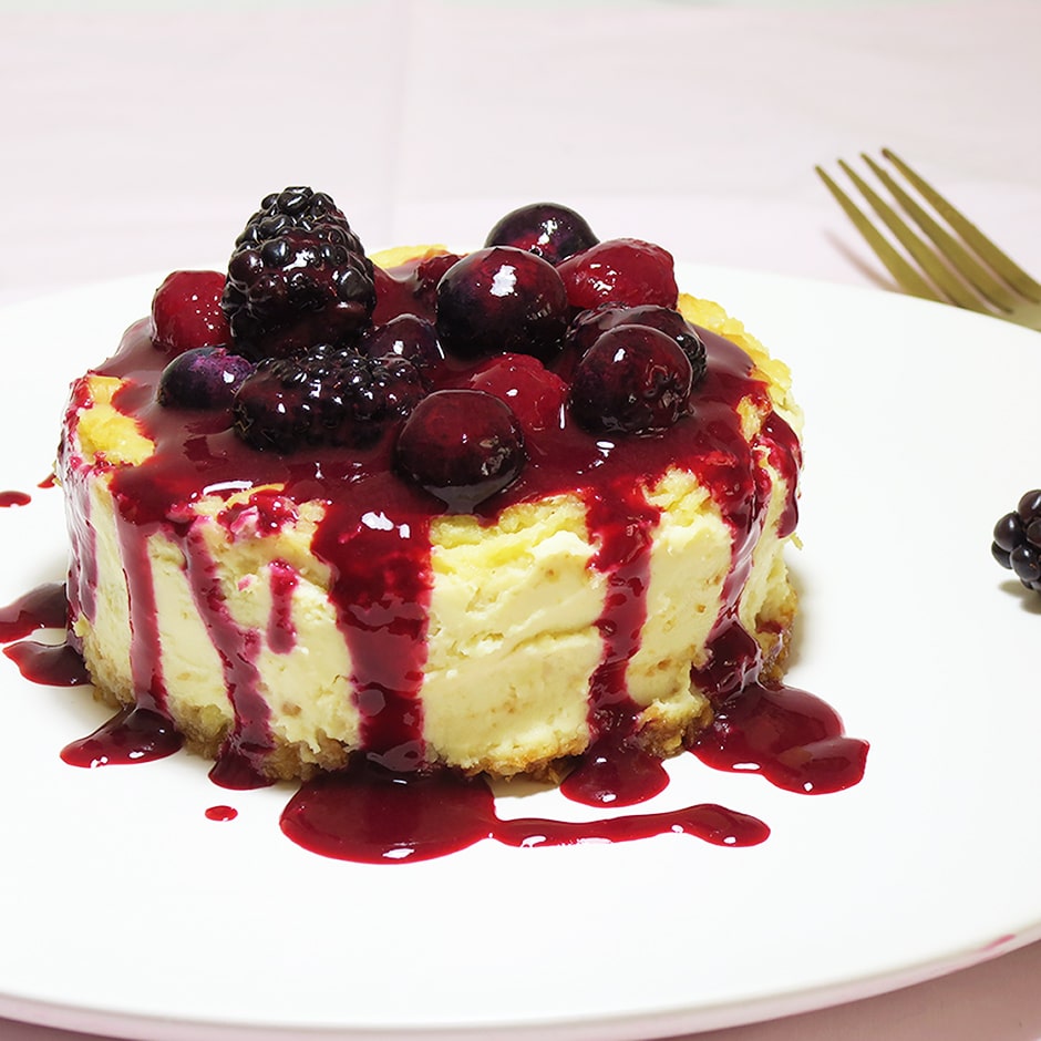 Baked lemon cheesecake topped with cherries, blackberries and blueberries. All on a white plate with a gold fork in the background.