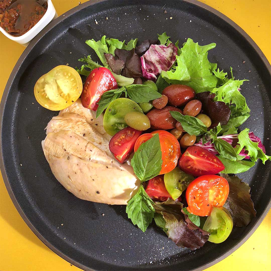 Mediterranean Salad featuring chicken breast, colourful tomatoes, different coloured olives, greens, and fresh basil, all on a black plate