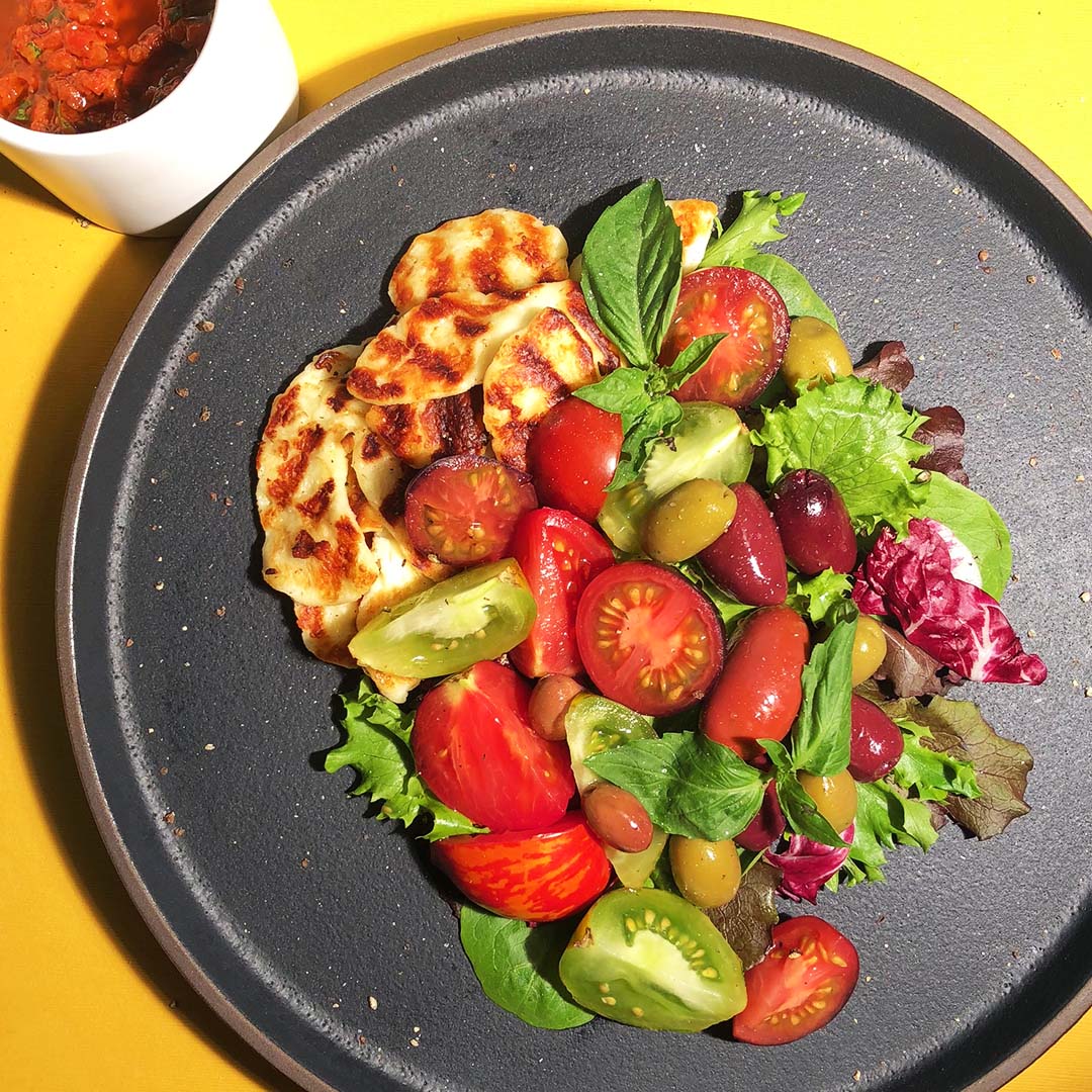 Mediterranean Salad, an entree salad with either chicken or Halloumi, tomatoes, olives, fresh basil, and vibrant greens, on a black plate