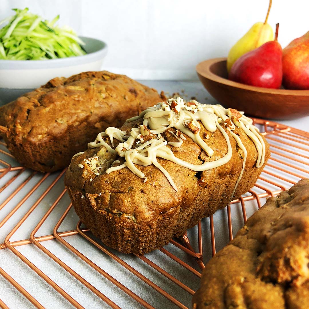 Three loaves of Pear and Zucchini Bread on a copper-coloured cooling rack, with a bowl of pears, and another bowl of shredded zucchini in the background.
