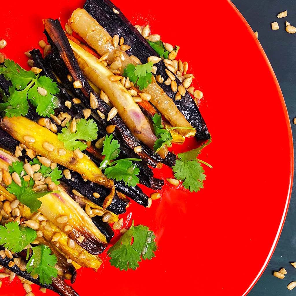Roasted rainbow carrots, sprinkled with sunflower seeds and cilantro, on a red plate.