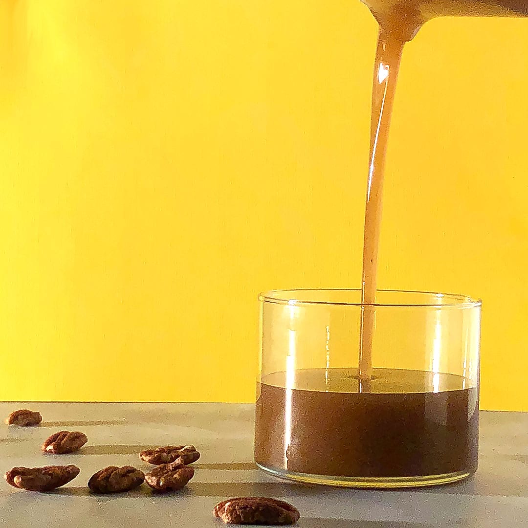 Easy vegan brown butter being poured into a glass container, against a yellow background, with pecans scattered nearby