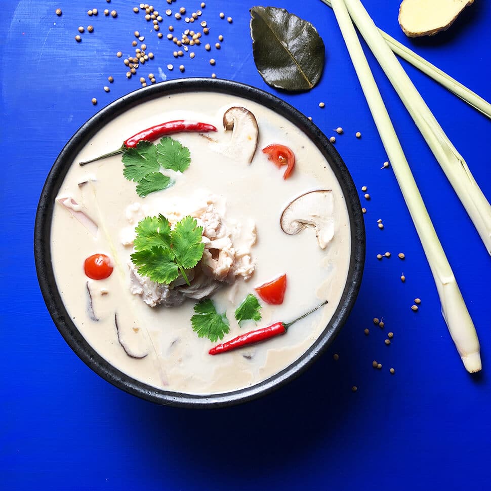 Tom Kha Soup with chicken in a black bowl on a bright blue background with coriander, lemongrass, ginger, and lime leaves.