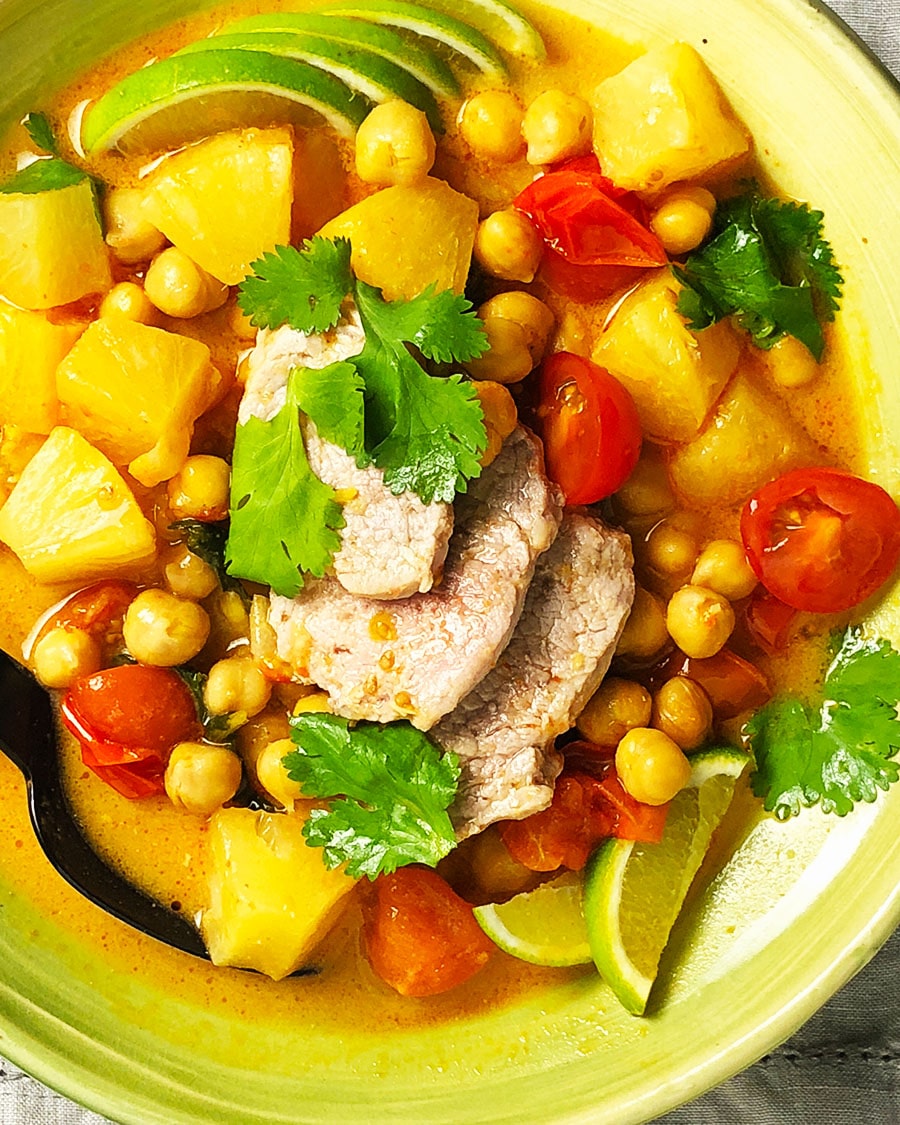 A close-up, topdown view of a bowl of Thai Chickpea Curry with Pork, containing red curry paste, coconut milk, chickpeas, tomatoes, pineapple, and seasonings.