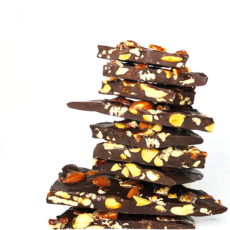 A towering pile of dark chocolate bark studded with pecans and almonds against a white background.
