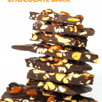 A towering pile of dark chocolate bark studded with pecans and almonds against a white background.
