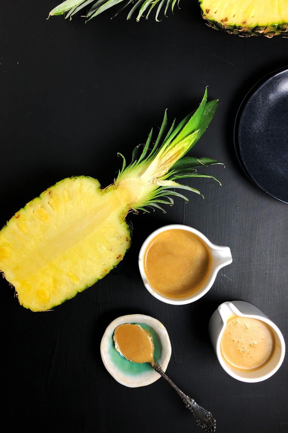 Top down view of two white vessels of Pineapple Brown Butter Teriyaki Sauce, alongside a spoon with some sauce, orange slices, and a halved pineapple, all against a black background.
