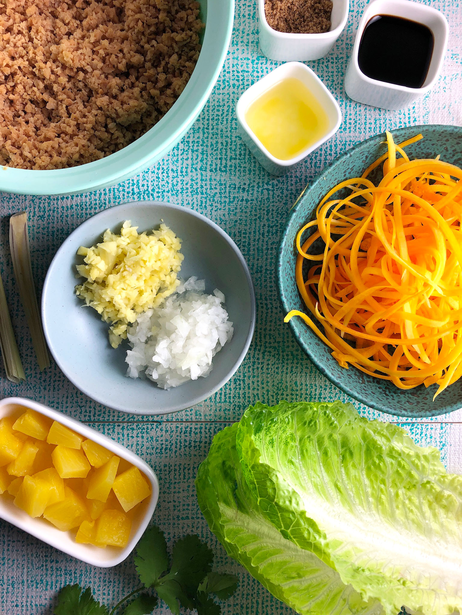 Top-down view of ingredients for lettuce wraps: Textured veggie protein (or pork), brown sugar, soya sauce, pineapple juice, carrots, lettuce, pineapple, diced onion, diced ginger