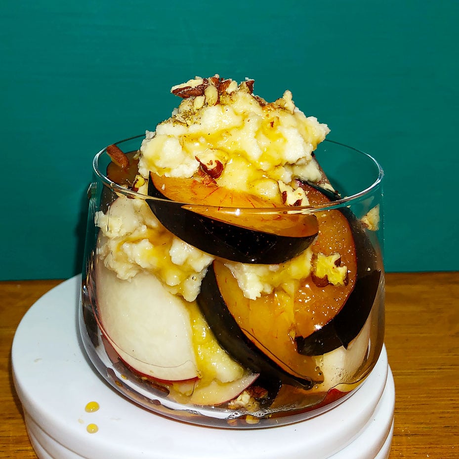 Side view of glass dish containing peaches, plums, dairy-free ricotta, pecans, and maple syrup