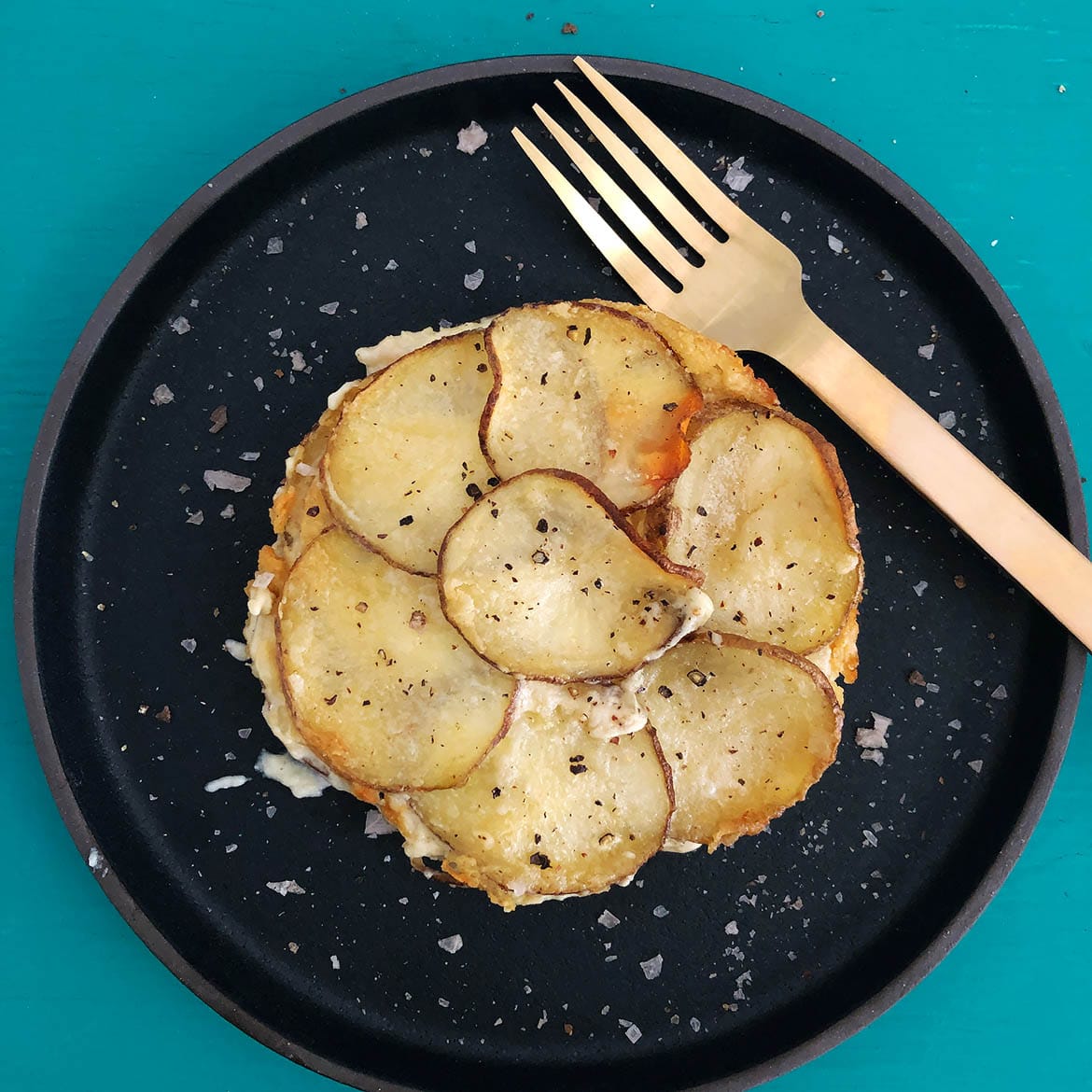 Top-down view of scalloped potatoes on a black plate with a gold fork