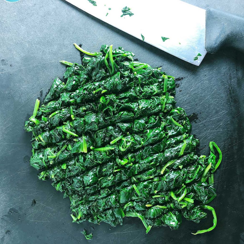 Top-down view of cooked, chopped spinach on a black cutting board, with knife
