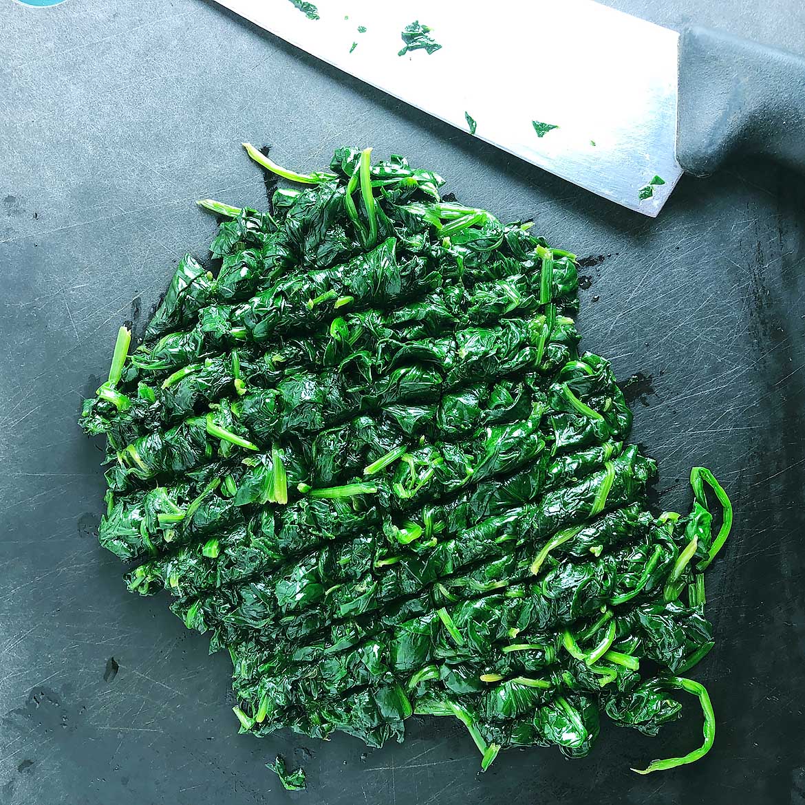 Top-down view of cooked, chopped spinach on a black cutting board, with knife