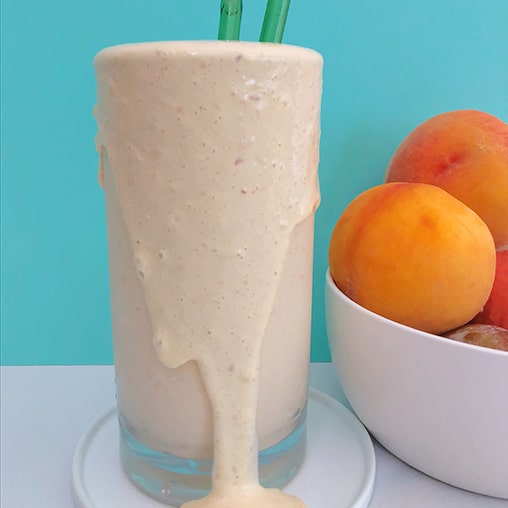 Double Peach Milkshake showing side view of peach milkshake pouring over the edge of a tall glass, with a bowl of peaches next to it. 