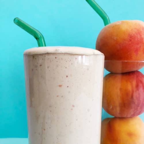 Side view of peach milkshake with a green glass straw, next to a glass of three peaches with a green glass straw, against a blue background