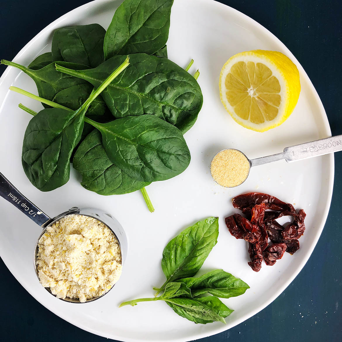 Top-down view of Hot Spinach Dip ingredients on a white plate: spinach, lemon, garlic powder, sun-dried tomatoes, basil, Mother Mix