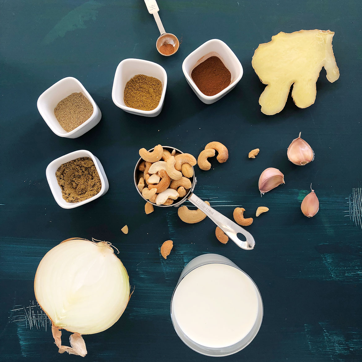 Top-down view of ingredients for Indian-inspired cashew sauce: onion, fresh ginger, garlic, garam masala, coriander, cardamom, cinnamon, and cashews