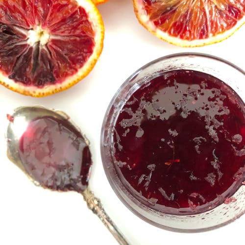 Blood Orange Syrup in a glass container, with cut halves of blood oranges in the background and a spoon with the syrup