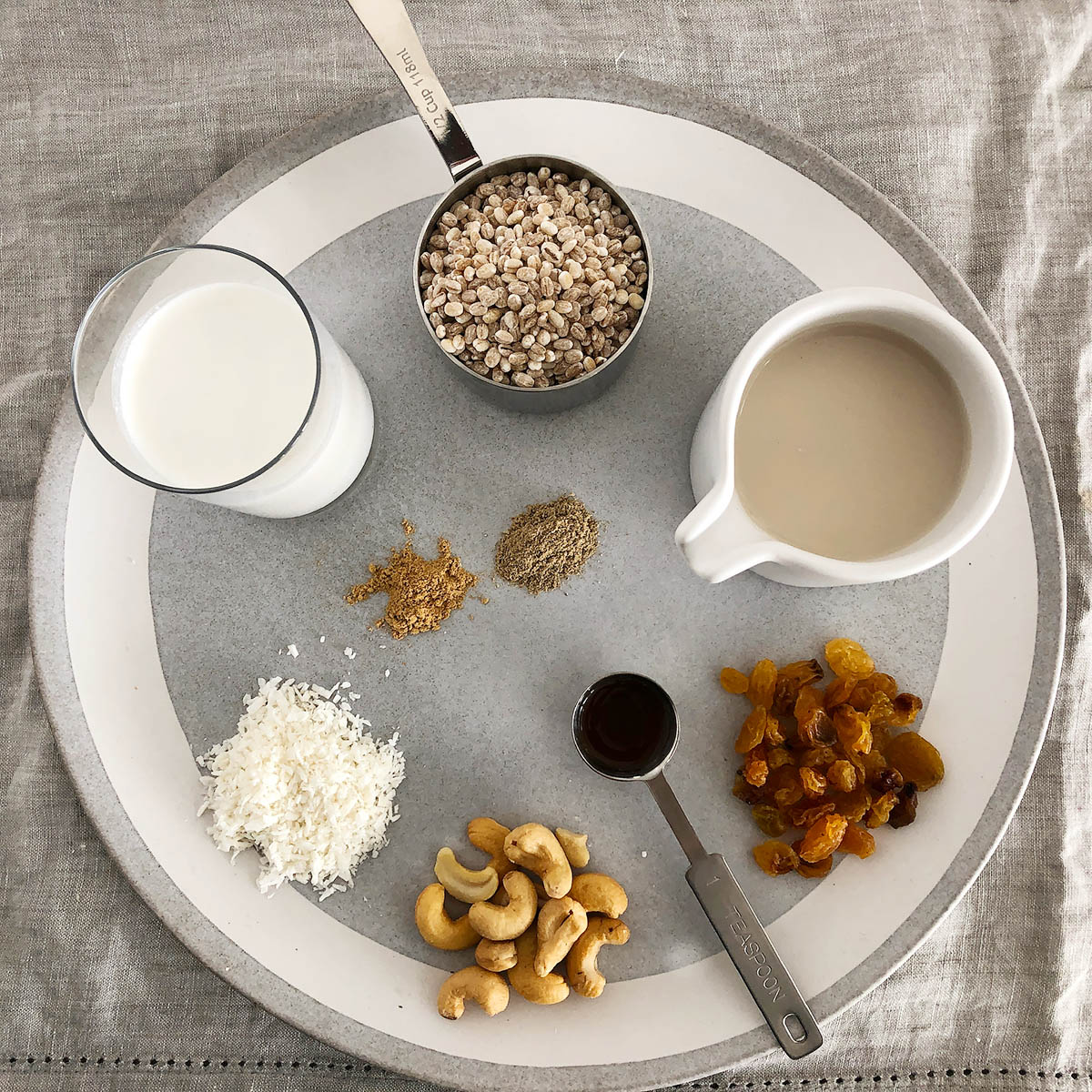 Ingredients for 1-Pot Barley Porridge: a measuring cup with pot barley, a vessel with cashew milk, a pile of raisins, a teaspoon of vanilla, cashews, shredded coconut, coconut milk, ginger, cardamom, all on a grey plate.