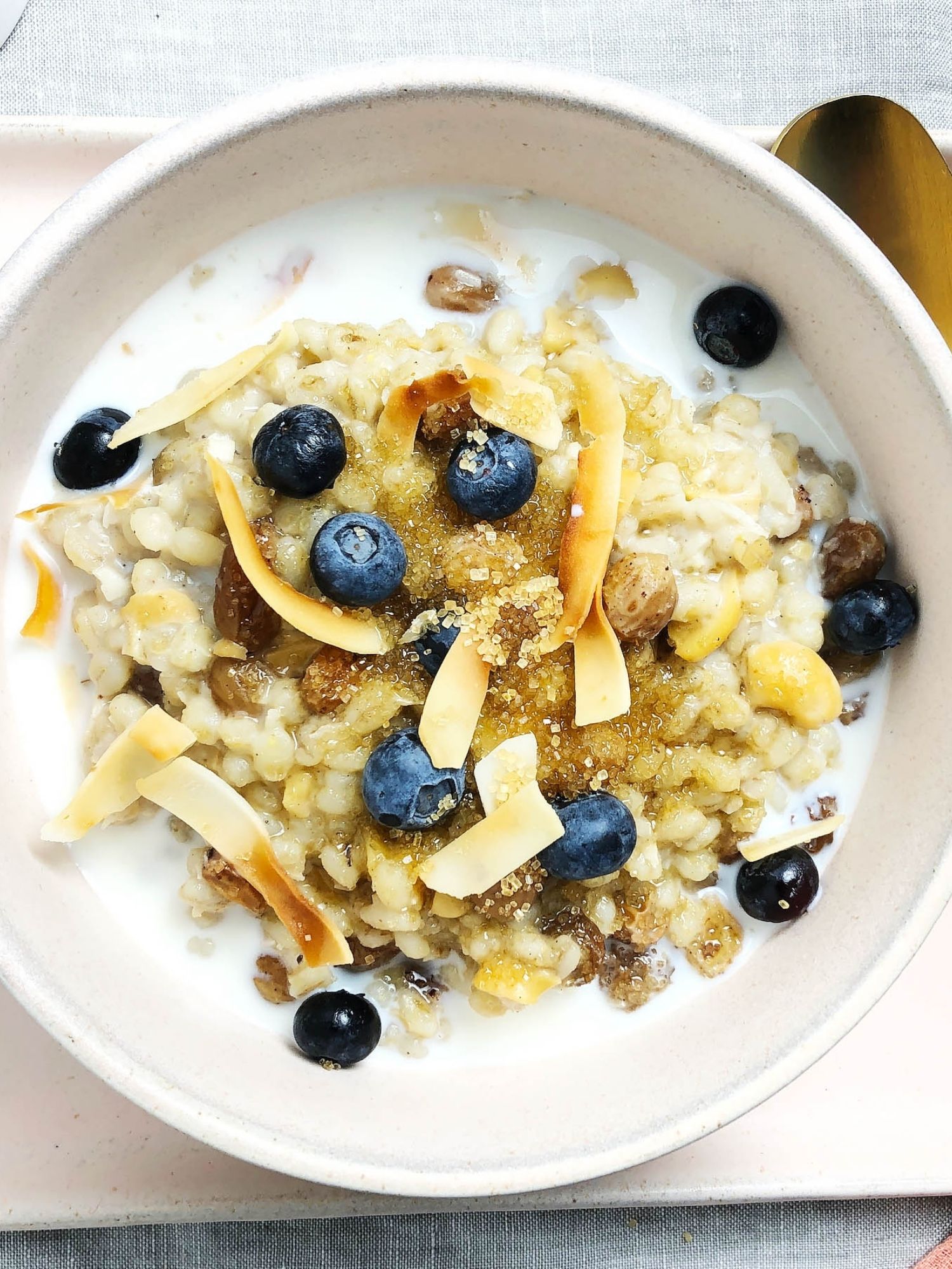 Top-down view of 1-Pot Barley Porridge in a light pink bowl. Porridge has been topped with toasted shredded coconut, fresh blueberries, and raw sugar.