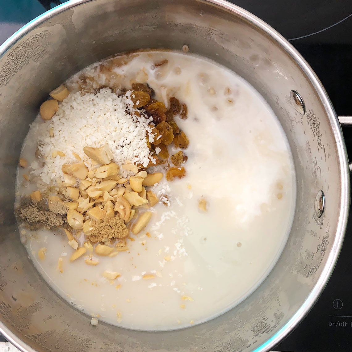 Top-down view of ingredients added to the cooked barley. These are cashew milk, coconut milk, chopped cashews, shredded coconut, raisins, vanilla, ginger, and cardamom. This is step 2 for making 1-Pot Barley Porridge.