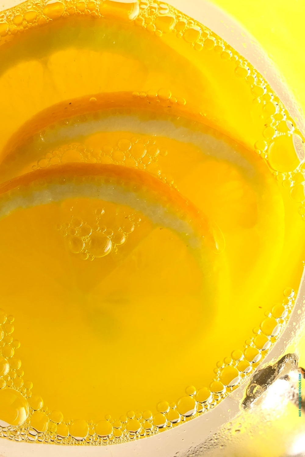 Top-down close up photo of hot lemonade bubbling in a clear glass. There are 3 lemon slices in the glass. The glass is set against a bright yellow background.