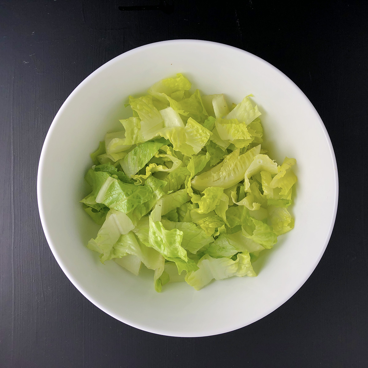 Top-down shot of chopped romaine lettuce in a white bowl, against a black background. Prep step number 1 for making Beyond Beef Taco Salad