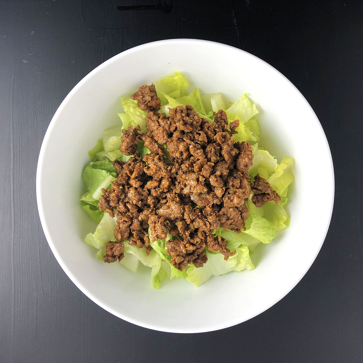 Top-down shot of chopped romaine lettuce and Beyond Beef Taco meat mix in a white bowl, against a black background. Prep step number 2 for making Beyond Beef Taco Salad