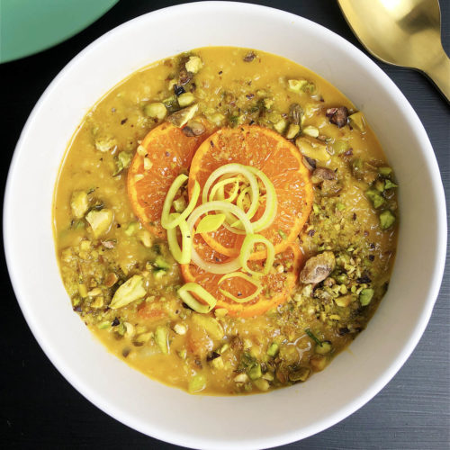 Top-down close up image of white bowl of lentil soup. Soup is garnished with pistachios, orange slices, and leek slices.