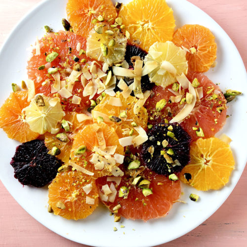 Top down shot of Simple Citrus Salad – slices of pink grapefruit, navel oranges, tangelos, blood oranges, and lemon, garnished with ribbons of toasted coconut, crushed pistachios and honey.