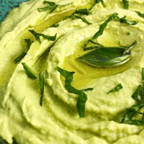 Top-down view of Edamame Hummus in a blue-speckled bowl. Hummus is pale green thanks to the basil and edamame beans. Hummus is topped with a bit of olive oil and fresh basil leaves.