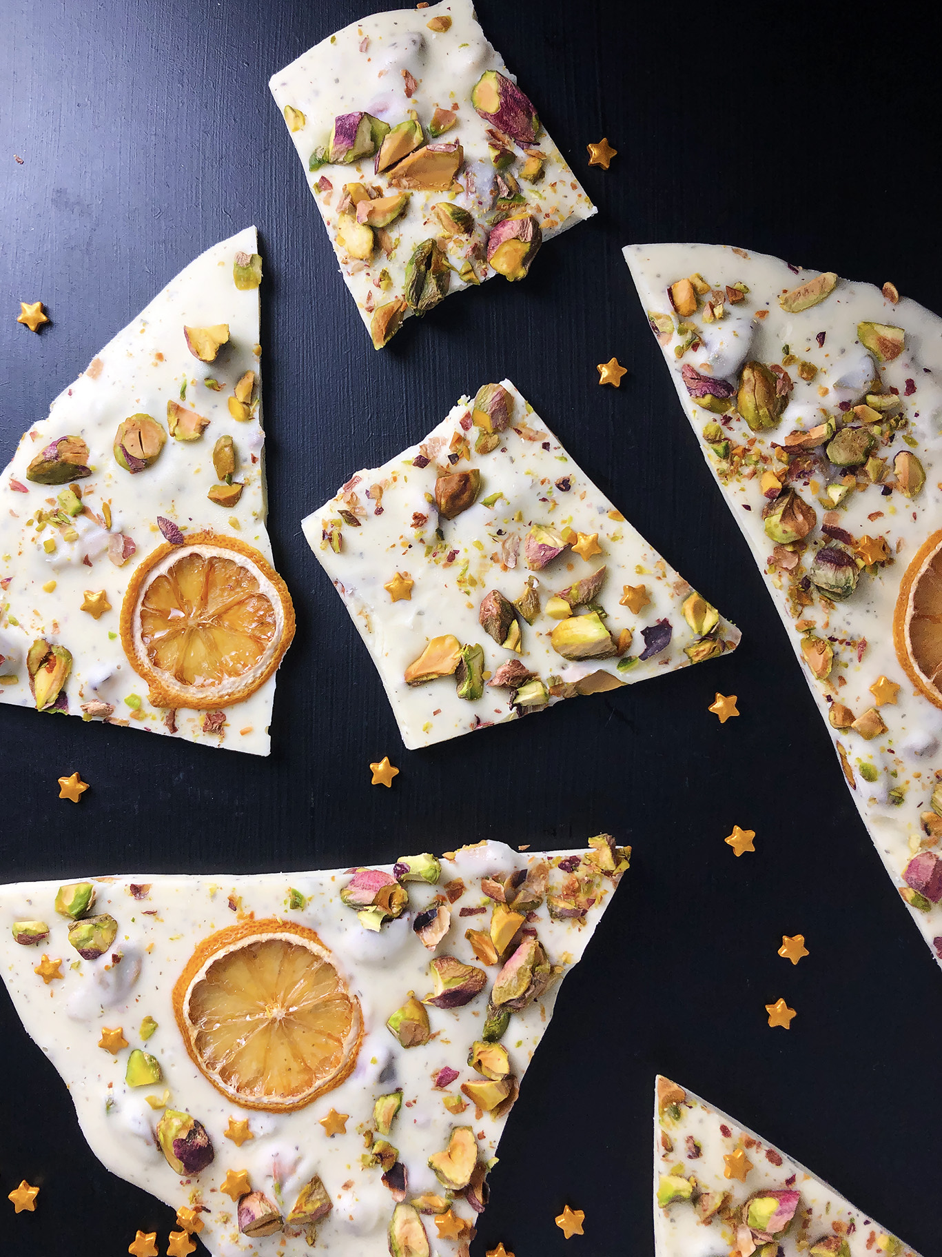 Top-down shot of white chocolate bark with lemon, green tea, pistachios and oven-dried lemons, against a black background, sprinkled with gold candy stars.