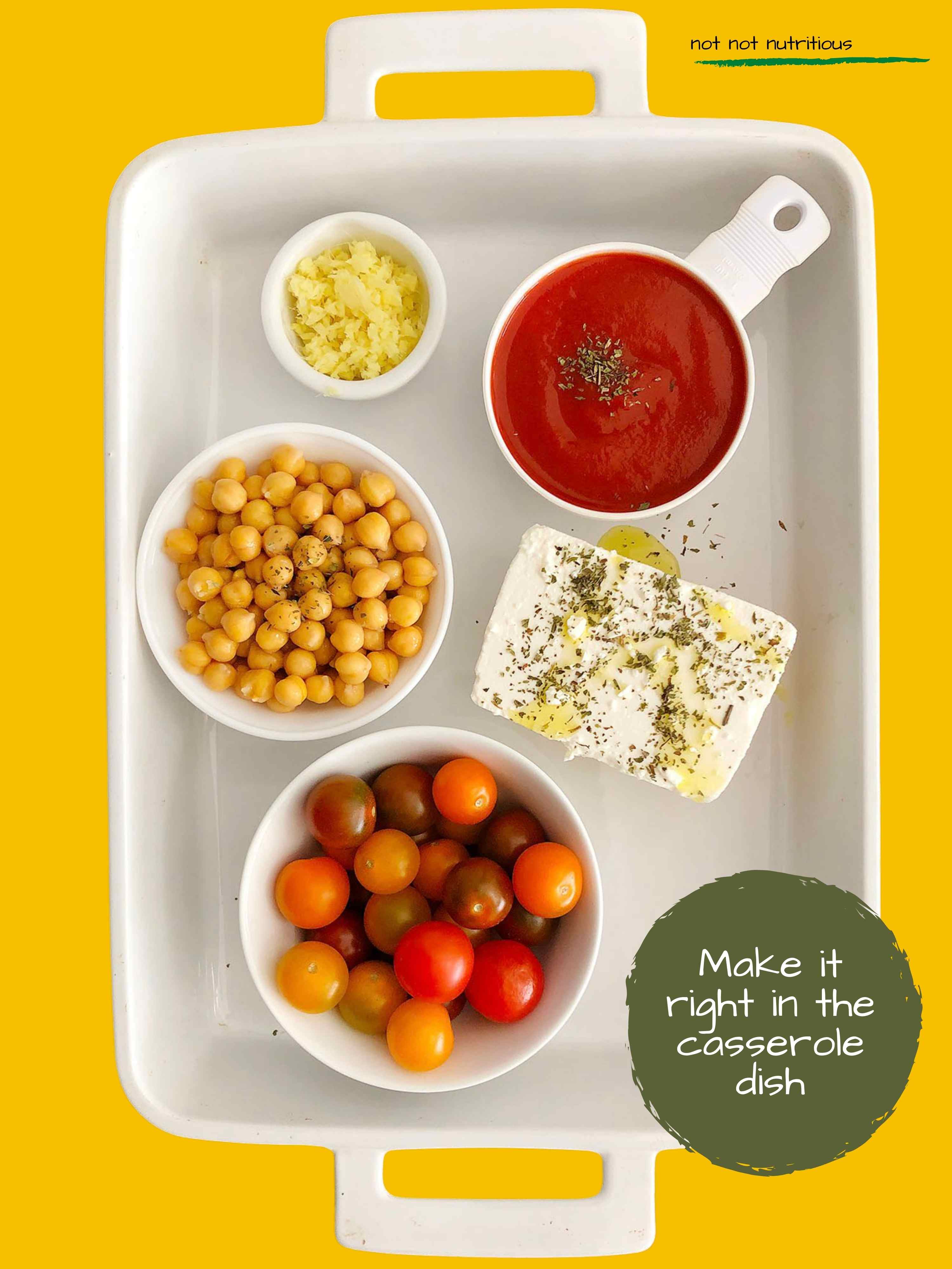 Top-down view of ingredients for Chickpea Feta Bake, assembled in a white casserole dish. From top right - tomato sauce, block of feta, tomatoes, chickpeas, minced ginger