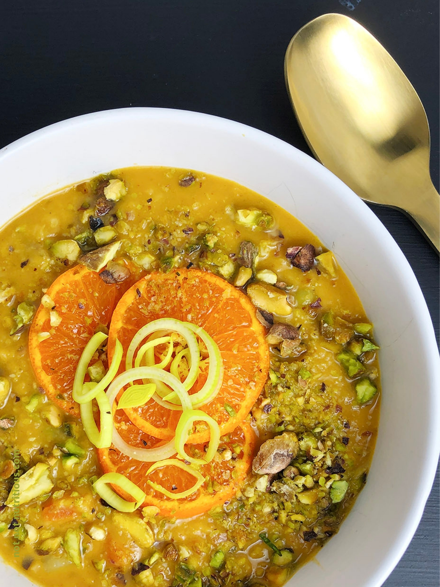 Photo is top-down close up image of white bowl of lentil soup. Soup is garnished with pistachios, orange slices, and leek slices.