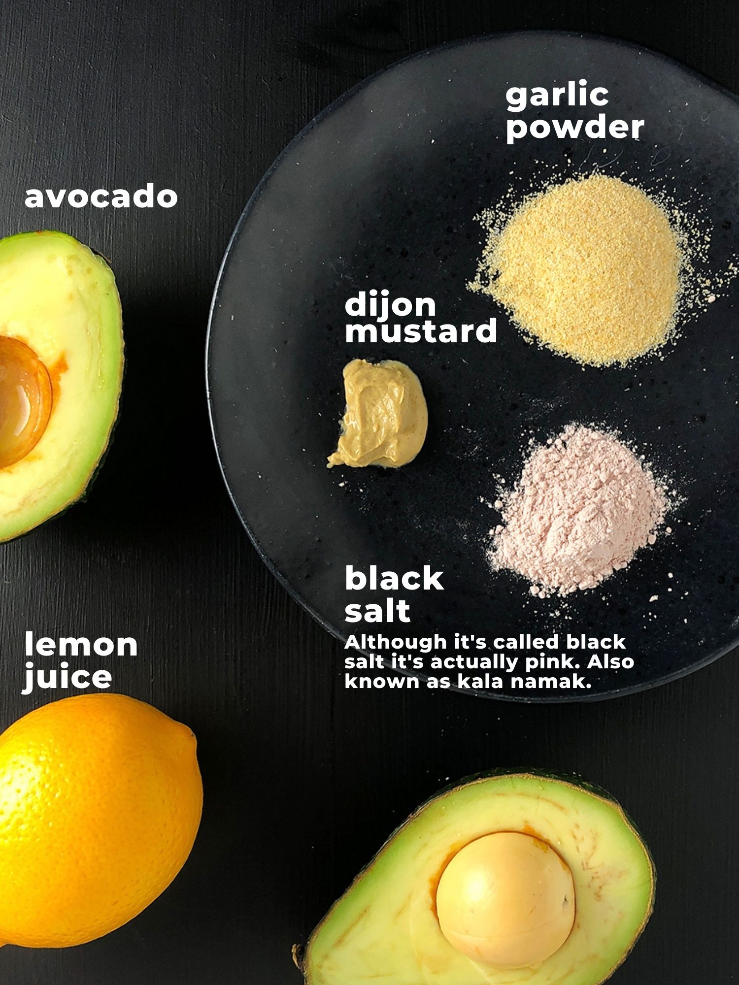 Infographic with a photograph of all the ingredients in Lemony Avocado Casesar Salad Dressing. Top-down shot shows half an avocado and a lemon, along with small amounts of garlic powder, dijon mustard, and black salt on a black plate. Text underneath black salt reads: Although it's called black salt it's actually pink. Also know as kala namak, it adds an eggy flavour, similar to traditional Caesar dressing. 
