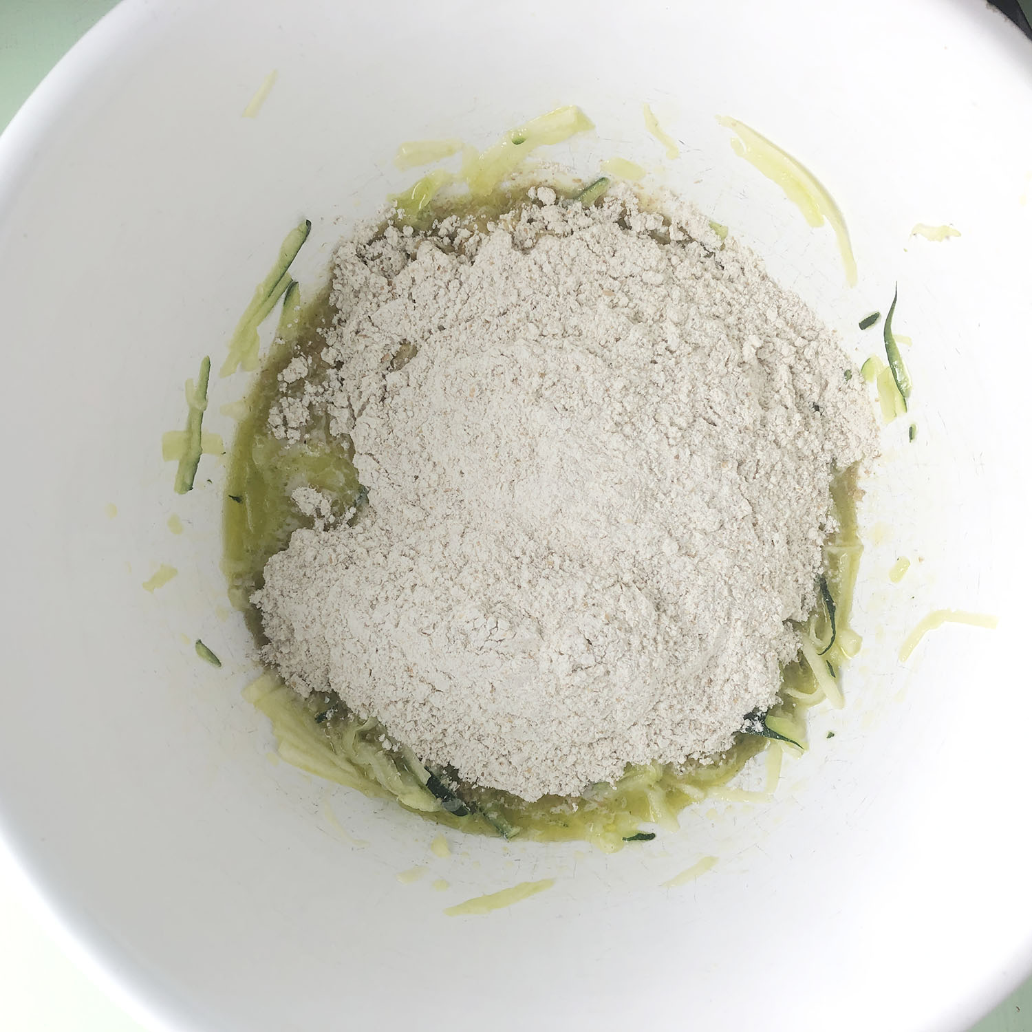 Top-down view of white bowl with zucchini, wet ingredients, flour, baking powder and salt.