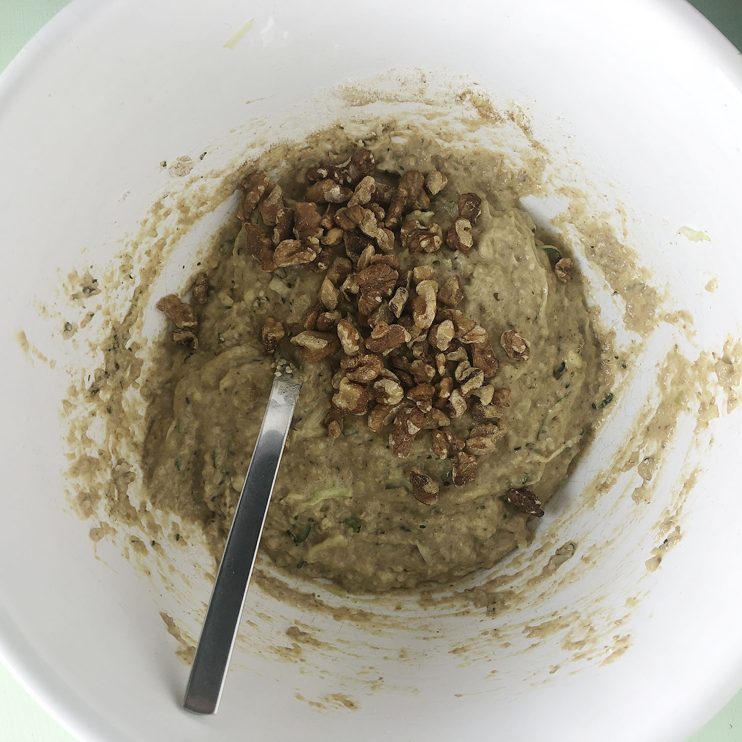 Top-down view of white mixing bowl with walnuts, zucchini and other ingredients for making Zucchini Chocolate Chunk Muffins