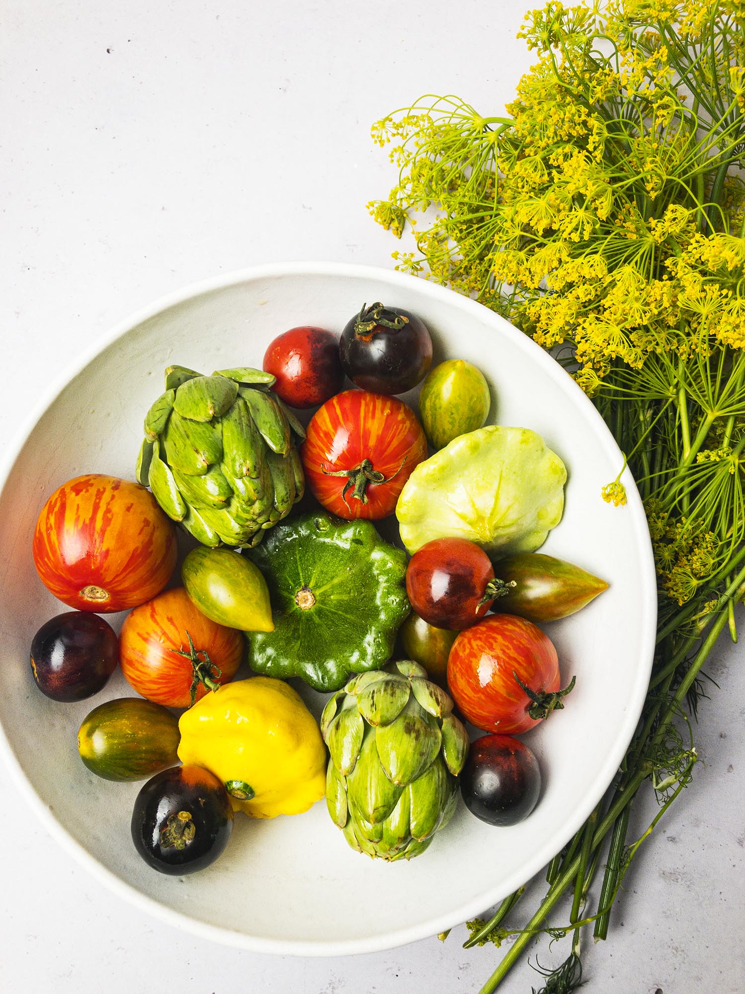 Vegetables in a white bowl: heirloom tomatoes, artichoke, and zucchini. A bouquet of dill surrounds the bowl.