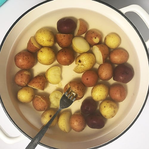 Top-down view of baby potatoes in a white pot, with a fork testing whether one of the potatoes is cooked 