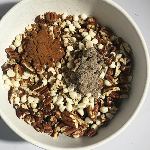 Top-down view of white mixing bowl with white chocolate chips, chopped pecans, cinnamon, cardamon, and coconut oil