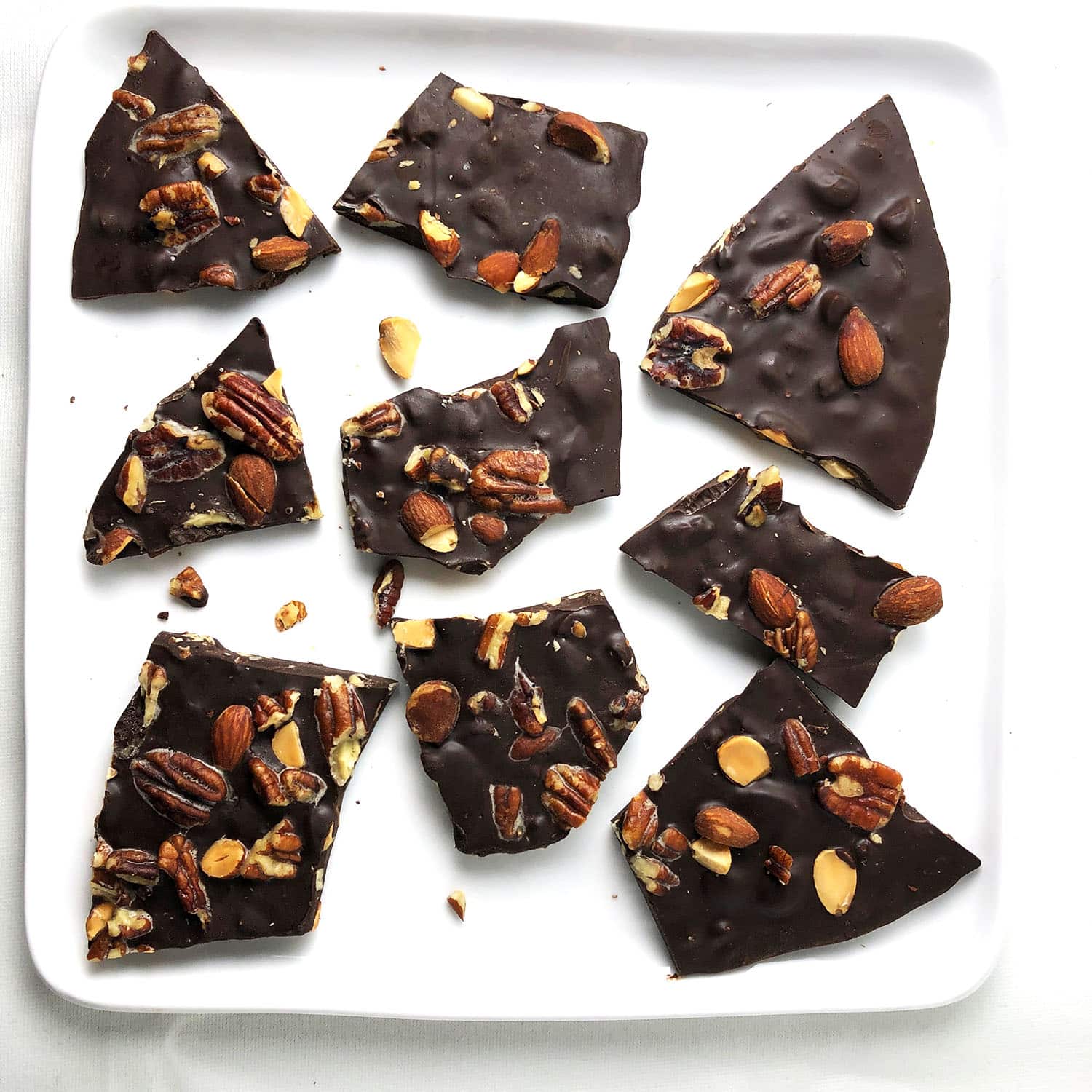 Top-down view of dark chocolate bark studded with buttery pecans and smoked almonds, on a square white plate.