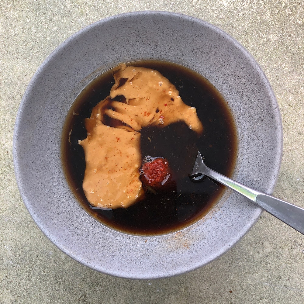 Top-down view of bowl with ingredients for Spicy Thai-Inspired Peanut Sauce, including peanut butter, coconut water, orange juice, soy sauce, black bean sauce, and coconut sugar. 