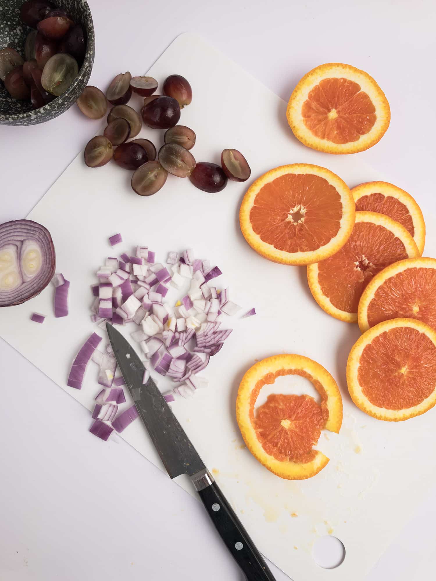 Top-down view of sliced oranges, sliced grapes, and red onions with a knife against a white background. 