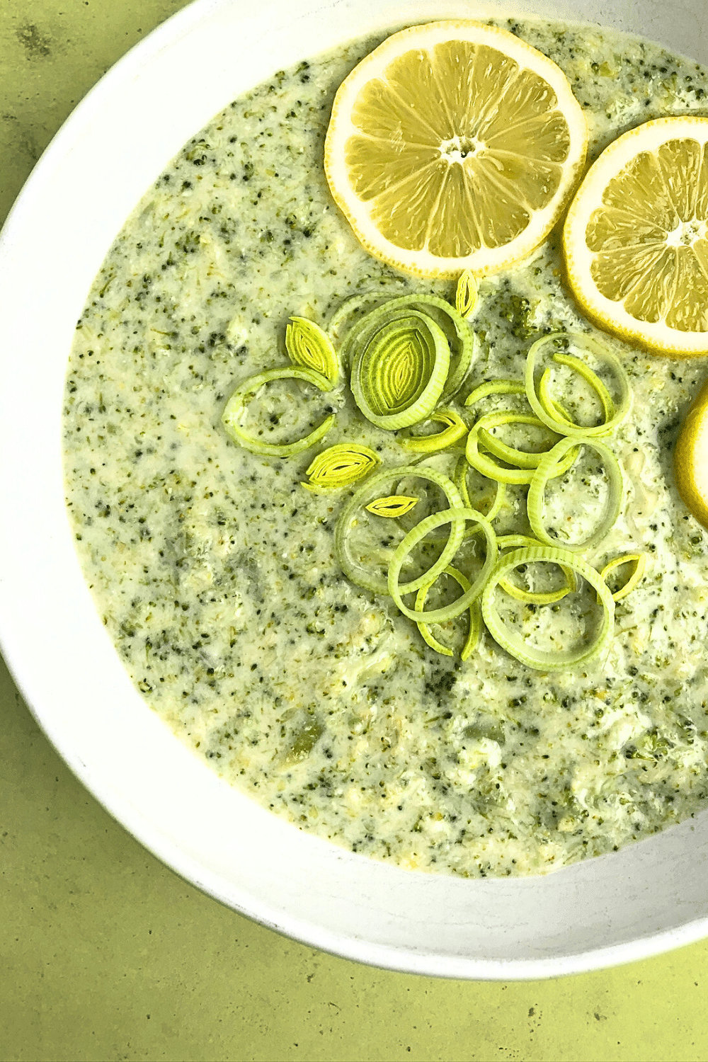 Top-down close up view of Creamy Lemon Leek and Broccoli Soup. The soup is in a white bowl and it is decorated with lemon slices and leek rings.