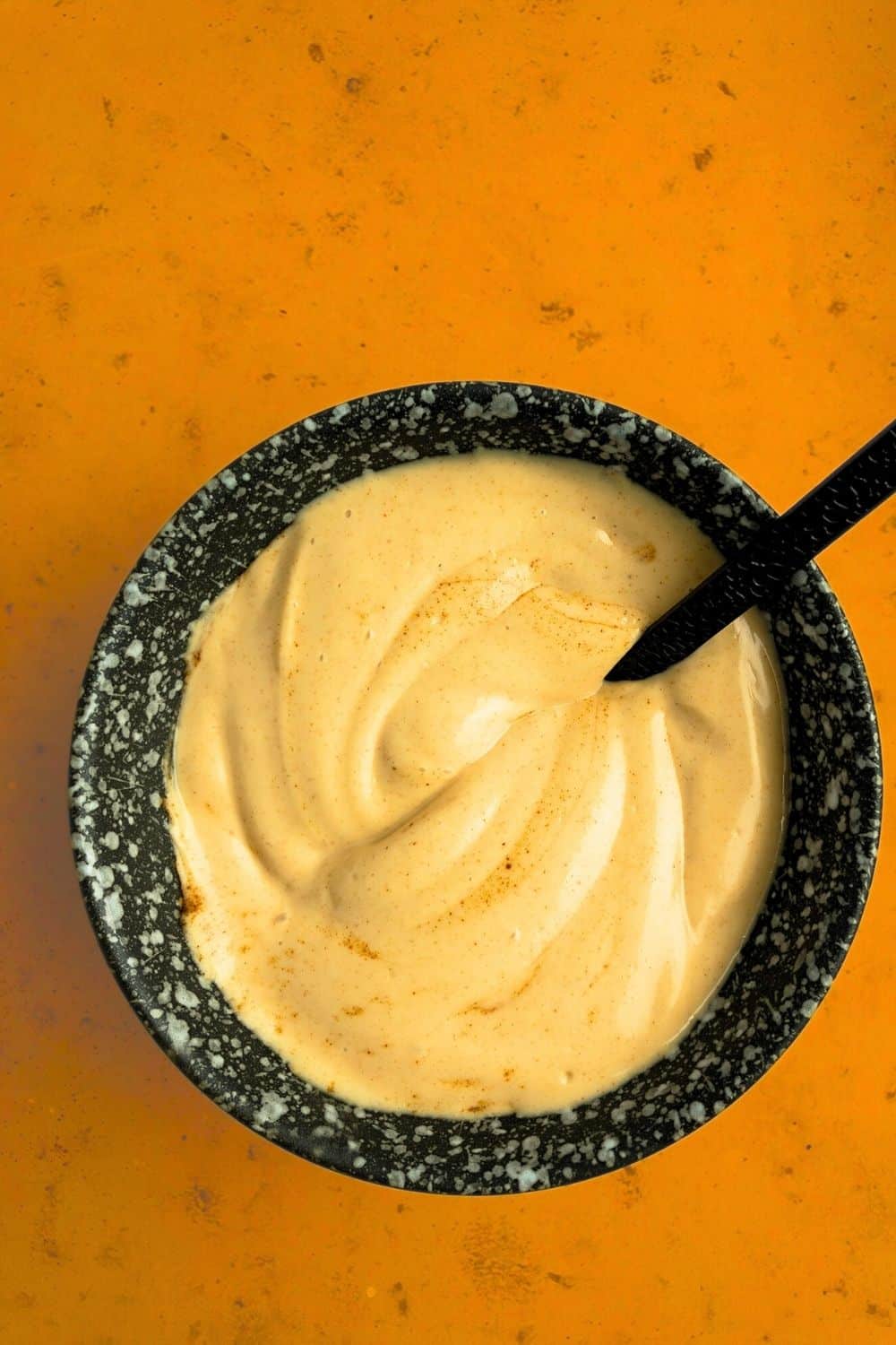 Top-down view of Smoky Maple Bacon Mayonnaise, in black bowl against a dark orange stone background