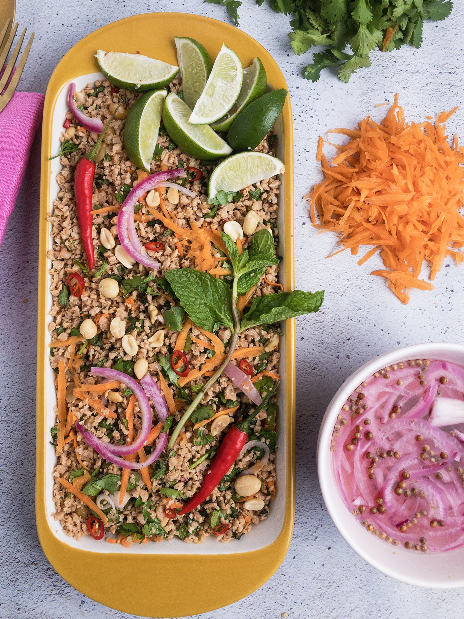 Pin for Thai-Inspired Vegan Larb, with top-down view of larb in a serving dish topped with limes, pickled shallots, carrots, and peanuts.
